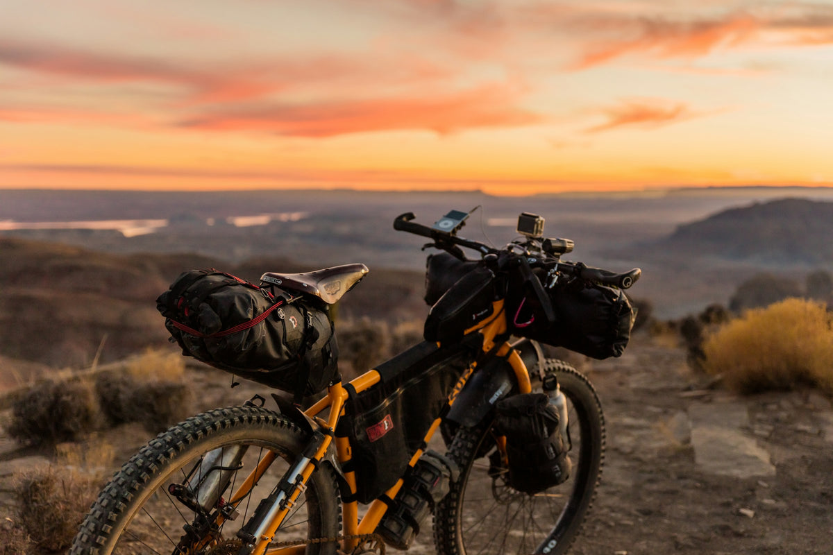 Voll beladenes Bikepacking-Rad mit Lenkerrolle und Satteltasche bei Sonnenuntergang über einem weiten Tal