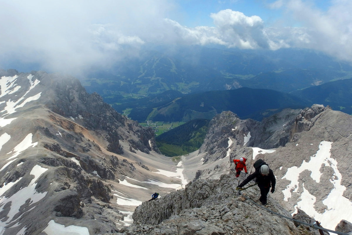 Zwei Bergsteiger mit Helm und Klettersteigausrüstung auf einem felsigen Grat, dahinter Alpenpanorama mit Schneefeldern