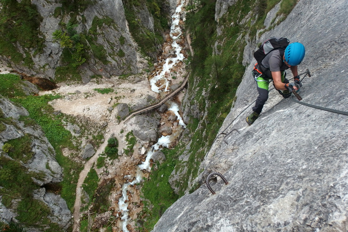 Kletterer mit Helm an einer steilen Felswand im Klettersteig, tief unten eine bewaldete Schlucht
