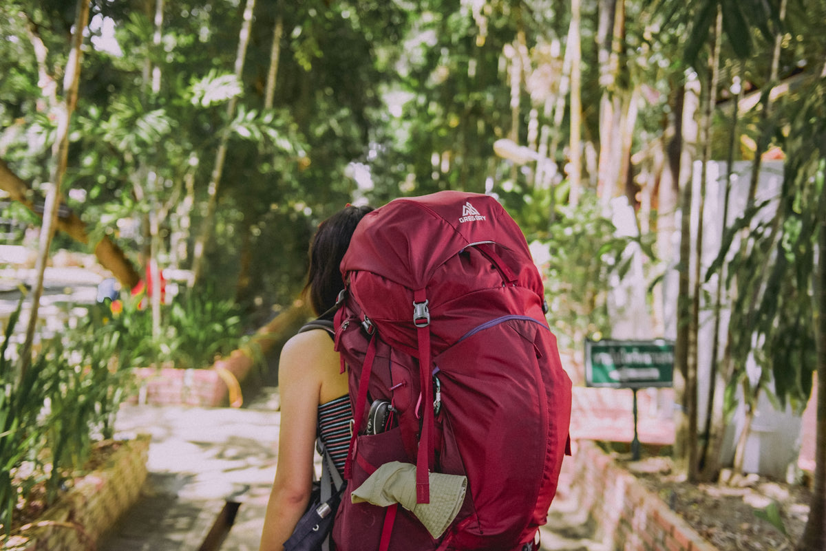Backpackerin mit großem roten Trekkingrucksack unterwegs auf einem grünen Pfad – Packliste Work & Travel