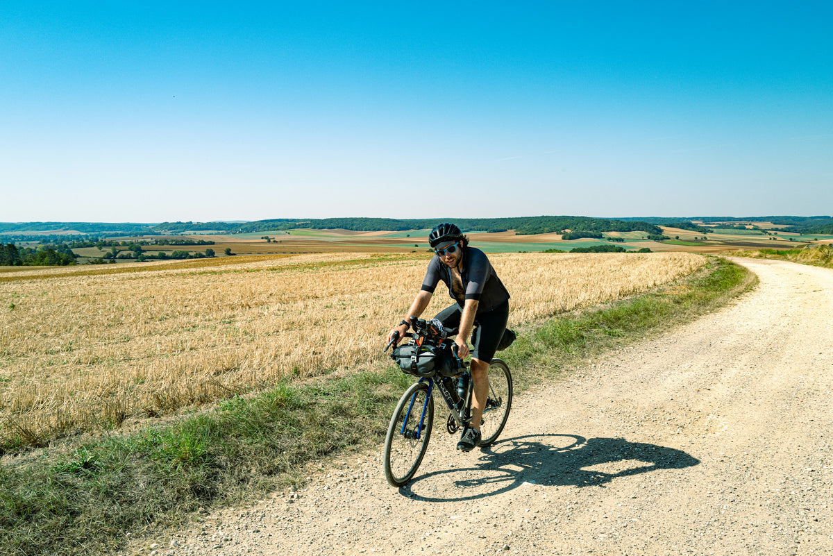Radfahrer mit Lenkertasche auf einem Schotterweg durch sommerliche Felder unter blauem Himmel