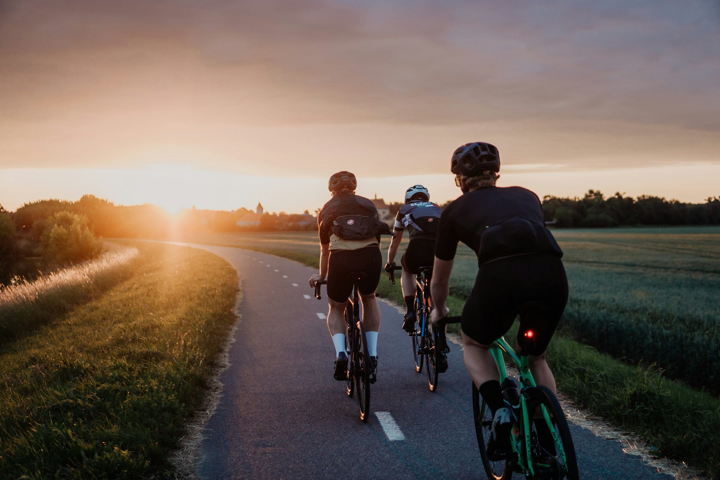 Rennradgruppe fährt bei Sonnenuntergang auf einer Landstraße zwischen Feldern